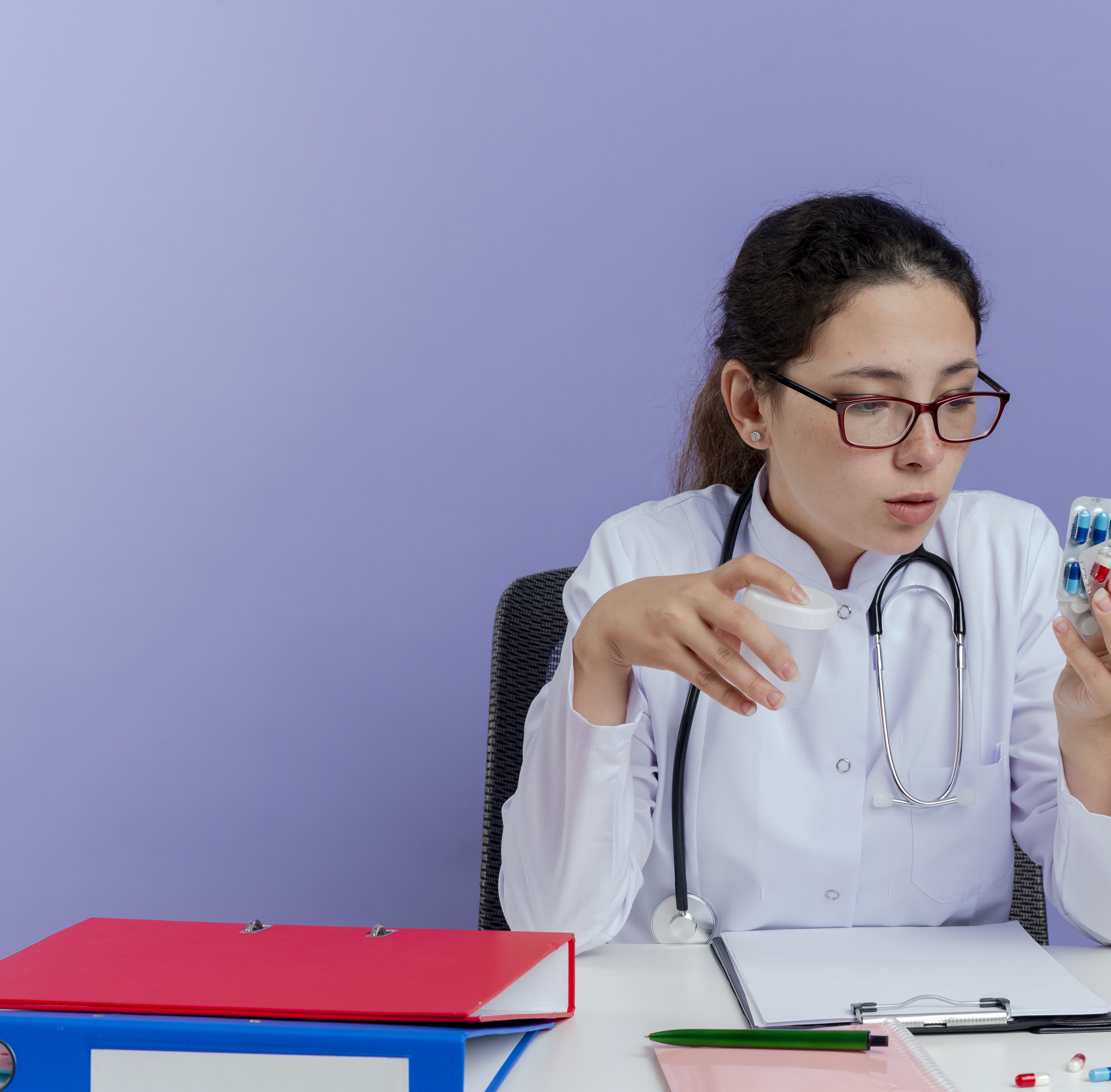 impressed-young-female-doctor-wearing-medical-robe-stethoscope-sitting-desk-with-medical-tools-holding-looking-medical-pills-holding-beaker-isolated